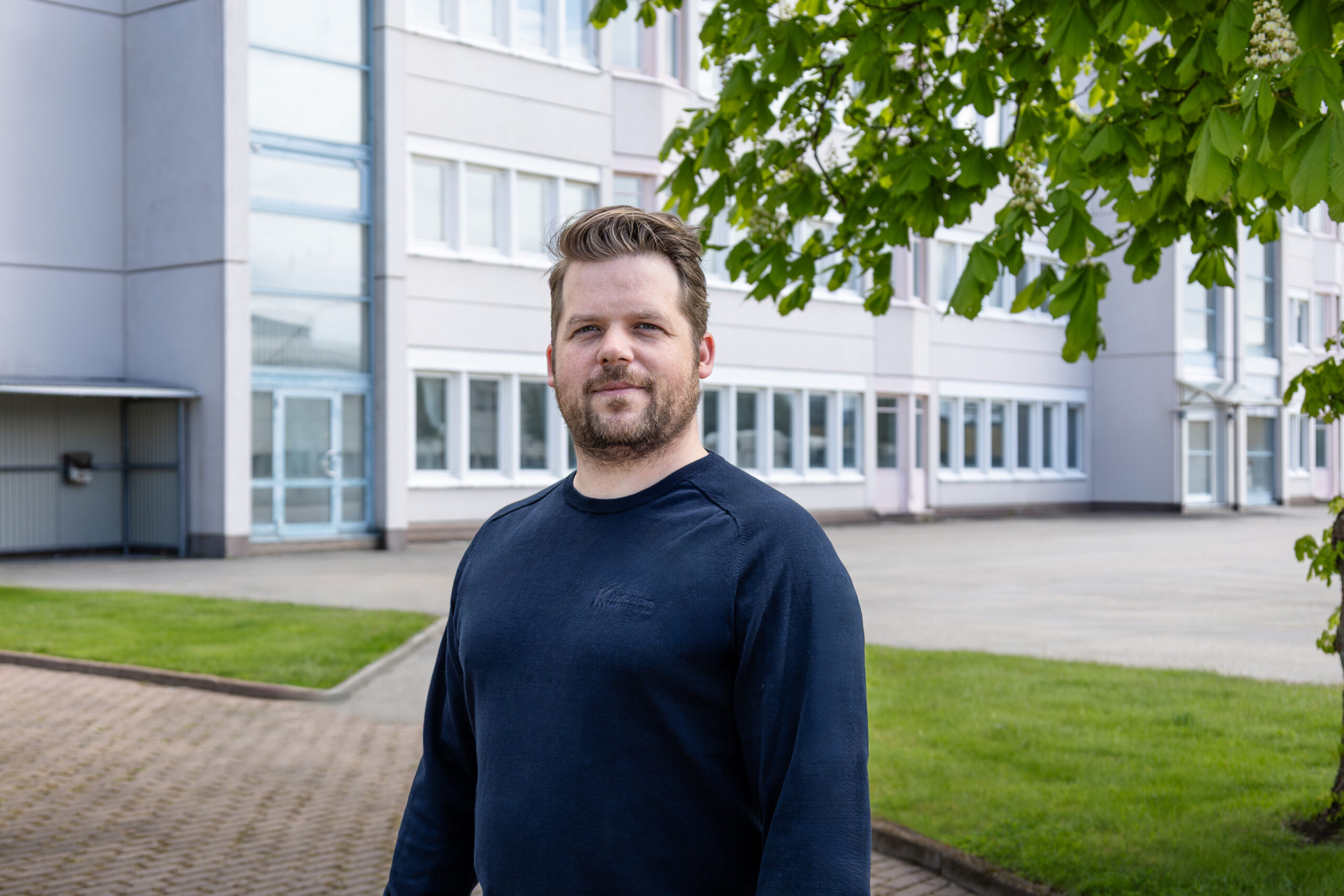 Kraftstadens uthyrare Marcus har på sig en marinblå tröja från Krafstaden. Han har skägg och kort hår. Byggnaden A01 syns bakom honom och löv från ett träd syns också i bakgrunden.